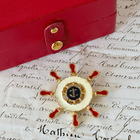Vintage Louis Giusti gold‑tone ship‑wheel brooch with white, red, and navy enamel and anchor motif, displayed on antique paper background.