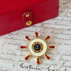 Vintage Louis Giusti gold‑tone ship‑wheel brooch with white, red, and navy enamel and anchor motif, displayed on antique paper background.
