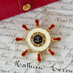 Vintage Louis Giusti gold‑tone ship‑wheel brooch with white, red, and navy enamel and anchor motif, displayed on antique paper background.