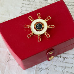 Vintage Louis Giusti gold‑tone ship‑wheel brooch with white, red, and navy enamel and anchor motif, displayed on antique paper background.