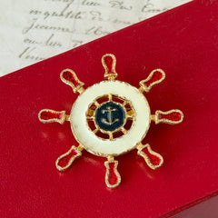 Vintage Louis Giusti gold‑tone ship‑wheel brooch with white, red, and navy enamel and anchor motif, displayed on antique paper background.