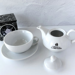 White porcelain teapot‑and‑cup set from La Route des Comptoirs with the boat‑and‑tea‑leaf logo on each piece