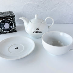 White porcelain teapot‑and‑cup set from La Route des Comptoirs with the boat‑and‑tea‑leaf logo on each piece