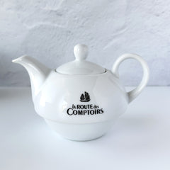 White porcelain teapot‑and‑cup set from La Route des Comptoirs with the boat‑and‑tea‑leaf logo on each piece