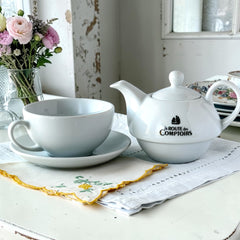 White porcelain teapot‑and‑cup set from La Route des Comptoirs with the boat‑and‑tea‑leaf logo on each piece