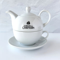 White porcelain teapot‑and‑cup set from La Route des Comptoirs with the boat‑and‑tea‑leaf logo on each piece