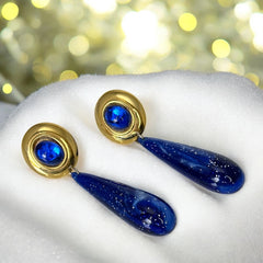 Vintage pierced earrings featuring glossy faux lapis long teardrop pendants and round blue cabochons set in gold-tone metal; likely acrylic components with cosmic speckled finish, measuring approx. 2¼" long, in very good vintage condition with minor glue visible on one cabochon edge.