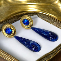 Vintage pierced earrings featuring glossy faux lapis long teardrop pendants and round blue cabochons set in gold-tone metal; likely acrylic components with cosmic speckled finish, measuring approx. 2¼" long, in very good vintage condition with minor glue visible on one cabochon edge.