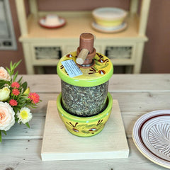 Hand-painted Provence herb mill with green, yellow, and brown olive branch motif on ceramic base, featuring a transparent chamber filled with dried herbs and topped with a wooden handle and cork stoppers.