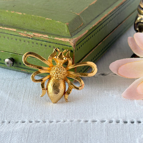 Vintage Mamselle gold‑tone bumblebee brooch with textured body and looped wings, signed on back, shown with floral and box props.