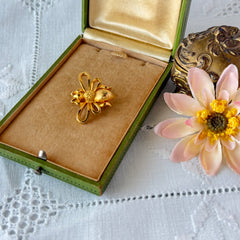Vintage Mamselle gold‑tone bumblebee brooch with textured body and looped wings, signed on back, shown with floral and box props.