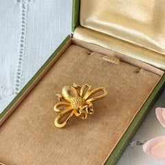 Vintage Mamselle gold‑tone bumblebee brooch with textured body and looped wings, signed on back, shown with floral and box props.
