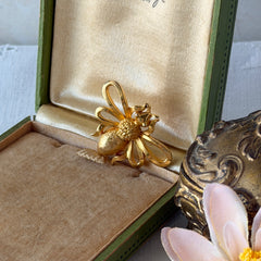 Vintage Mamselle gold‑tone bumblebee brooch with textured body and looped wings, signed on back, shown with floral and box props.