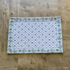 Reversible quilted cotton placemat in Lauris Light Green by Marat d’Avignon, featuring a white center with lavender purple floral medallions and a border of lavender sprigs and olive branches, with a white reverse side  scattered with tiny lavender  blossoms, crafted in Provence.