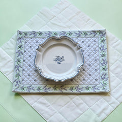 Reversible quilted cotton placemat in Lauris Light Green by Marat d’Avignon, featuring a white center with lavender purple floral medallions and a border of lavender sprigs and olive branches, with a white reverse side  scattered with tiny lavender  blossoms, crafted in Provence.