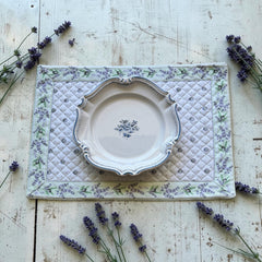 Reversible quilted cotton placemat in Lauris Light Green by Marat d’Avignon, featuring a white center with lavender purple floral medallions and a border of lavender sprigs and olive branches, with a white reverse side  scattered with tiny lavender  blossoms, crafted in Provence.