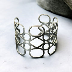 Silver tone cuff bracelet with polished openwork design featuring interconnected ovals, circles, and diamond shapes; modern, unsigned piece with a sleek finish and subtle surface wear.