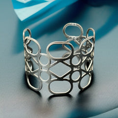 Silver tone cuff bracelet with polished openwork design featuring interconnected ovals, circles, and diamond shapes; modern, unsigned piece with a sleek finish and subtle surface wear.