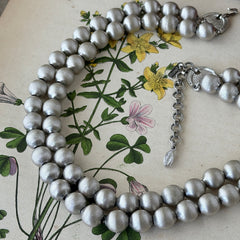 Vintage Monet brushed silver tone two‑strand bead necklace displayed on a black velvet bust and vintage botanical background.