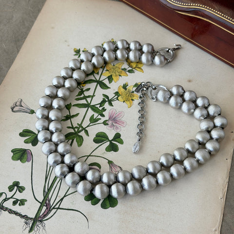 Vintage Monet brushed silver tone two‑strand bead necklace displayed on a black velvet bust and vintage botanical background.