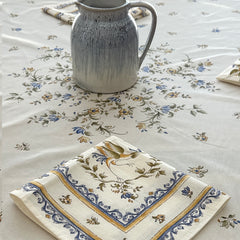 Round coated cotton tablecloth in white with polychrome and blue Provençal motifs, featuring birds, butterflies, and floral designs inspired by traditional Moustiers faience
