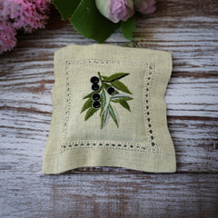 Ecru cotton sachet pillow embroidered with a branch of black olives and green leaves, framed by a lace-like hemstitch border. Filled with fragrant French lavender buds, this handmade cushion evokes rustic Mediterranean charm and artisanal elegance.
