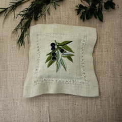 Ecru cotton sachet pillow embroidered with a branch of black olives and green leaves, framed by a lace-like hemstitch border. Filled with fragrant French lavender buds, this handmade cushion evokes rustic Mediterranean charm and artisanal elegance.