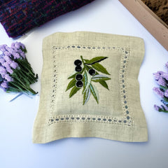 Ecru cotton sachet pillow embroidered with a branch of black olives and green leaves, framed by a lace-like hemstitch border. Filled with fragrant French lavender buds, this handmade cushion evokes rustic Mediterranean charm and artisanal elegance.