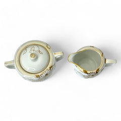 Vintage Nippon porcelain sugar bowl and creamer set with hand-painted gold floral design on white glaze; sugar bowl with dual handles and lid, creamer with spout and single handle; both marked “Hand Painted Nippon” with sun emblem on base.