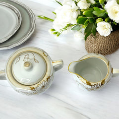 Vintage Nippon porcelain sugar bowl and creamer set with hand-painted gold floral design on white glaze; sugar bowl with dual handles and lid, creamer with spout and single handle; both marked “Hand Painted Nippon” with sun emblem on base.