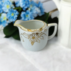 Vintage Nippon porcelain sugar bowl and creamer set with hand-painted gold floral design on white glaze; sugar bowl with dual handles and lid, creamer with spout and single handle; both marked “Hand Painted Nippon” with sun emblem on base.