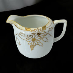 Vintage Nippon porcelain sugar bowl and creamer set with hand-painted gold floral design on white glaze; sugar bowl with dual handles and lid, creamer with spout and single handle; both marked “Hand Painted Nippon” with sun emblem on base.