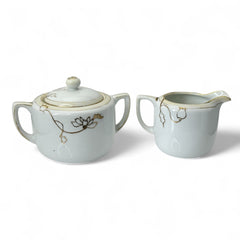 Vintage Nippon porcelain sugar bowl and creamer set with hand-painted gold floral design on white glaze; sugar bowl with dual handles and lid, creamer with spout and single handle; both marked “Hand Painted Nippon” with sun emblem on base.