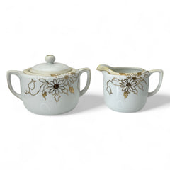 Vintage Nippon porcelain sugar bowl and creamer set with hand-painted gold floral design on white glaze; sugar bowl with dual handles and lid, creamer with spout and single handle; both marked “Hand Painted Nippon” with sun emblem on base.