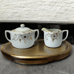 Vintage Nippon porcelain sugar bowl and creamer set with hand-painted gold floral design on white glaze; sugar bowl with dual handles and lid, creamer with spout and single handle; both marked “Hand Painted Nippon” with sun emblem on base.