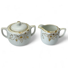 Vintage Nippon porcelain sugar bowl and creamer set with hand-painted gold floral design on white glaze; sugar bowl with dual handles and lid, creamer with spout and single handle; both marked “Hand Painted Nippon” with sun emblem on base.