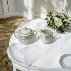 Vintage Nippon porcelain sugar bowl and creamer set with hand-painted gold floral design on white glaze; sugar bowl with dual handles and lid, creamer with spout and single handle; both marked “Hand Painted Nippon” with sun emblem on base.