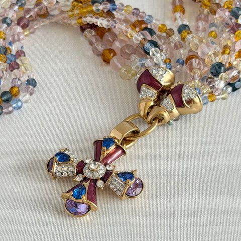 Vintage Nolan Miller necklace with an ornate enamel and rhinestone cross pendant on an eight‑strand pastel faceted glass bead torsade in gold‑tone metal.