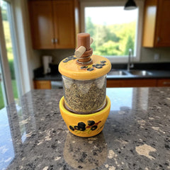 Provence hand-painted ceramic herb grinder with a yellow base featuring black olive and green leaf design, filled with dried Herbes de Provence. 