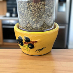 Provence hand-painted ceramic herb grinder with a yellow base featuring black olive and green leaf design, filled with dried Herbes de Provence. 