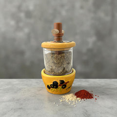 Provence hand-painted ceramic herb grinder with a yellow base featuring black olive and green leaf design, filled with dried Herbes de Provence. 