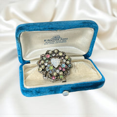 Vintage silver tone brooch with ornate round filigree design, featuring a central opalite heart cabochon and pastel rhinestones in pink, blue, yellow, and lavender—shown front and back to highlight detailing and clasp.