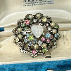 Vintage silver tone brooch with ornate round filigree design, featuring a central opalite heart cabochon and pastel rhinestones in pink, blue, yellow, and lavender—shown front and back to highlight detailing and clasp.