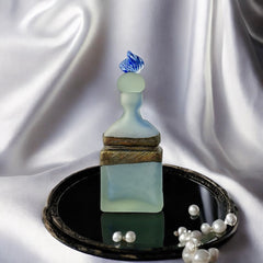 Vintage Romanian art glass perfume bottle from the 1990s, featuring frosted blue and green glass with a sculptural swirled stopper and bronze wire bow detail around the neck. Square base with a tapered neck; dauber slightly shortened due to breakage.