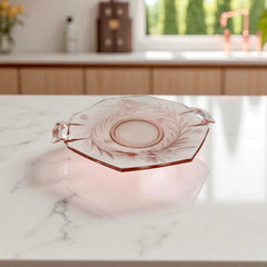 Vintage pink Depression glass serving dish with etched floral design and dual handles, styled on marble with soft lighting to highlight its shape, color, and collectible charm.