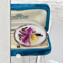 Vintage French reverse carved Lucite brooch featuring a hand-painted pink and yellow orchid on a white background. Oval shape with dimensional floral detail, excellent condition, circa 1950s.
