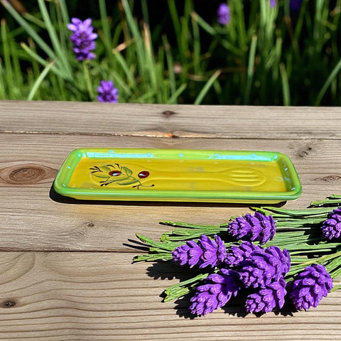Rectangular ceramic spoon rest with yellow interior and green polka-dot rim, featuring hand-painted olive leaves, red berries, and whimsical fork and spoon motif; measures 11 by 4 inches.