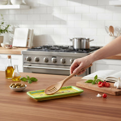 Rectangular ceramic spoon rest with yellow interior and green polka-dot rim, featuring hand-painted olive leaves, red berries, and whimsical fork and spoon motif; measures 11 by 4 inches.