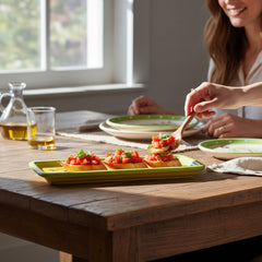 Rectangular ceramic spoon rest with yellow interior and green polka-dot rim, featuring hand-painted olive leaves, red berries, and whimsical fork and spoon motif; measures 11 by 4 inches.