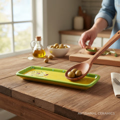 Rectangular ceramic spoon rest with yellow interior and green polka-dot rim, featuring hand-painted olive leaves, red berries, and whimsical fork and spoon motif; measures 11 by 4 inches.